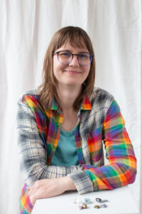 Cher Hamilton-Tekautz is a white woman with brown shoulder length hair wearing glasses and a rainbow plaid flannel. There are crystals laid on the stand she is resting on.  