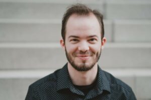 Aaron Melnek is a white man with combed back dark brown hair and facial hair, wearing a dark, collared shirt. 